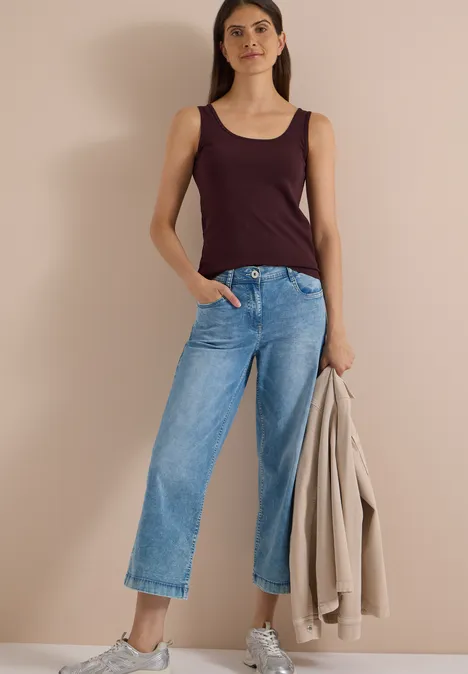 Vrouw in wijde jeans met bordeaux top, houdt een beige jas vast.