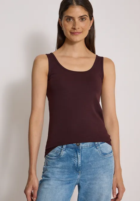 Vrouw staat voor neutrale achtergrond, draagt een bordeaux top en blauwe jeans.