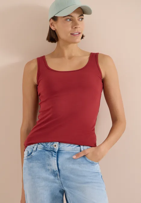 Femme portant un débardeur rouge et un jean bleu devant un mur beige, une main dans la poche.
