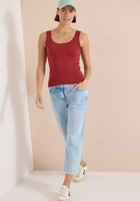 Femme en débardeur rouge, jean bleu clair et baskets blanches sur fond neutre.
