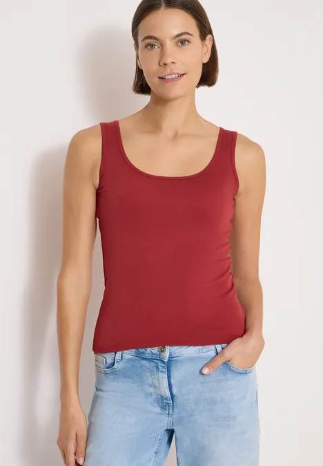 Femme en débardeur rouge et jean bleu clair devant un mur clair, une main dans la poche.