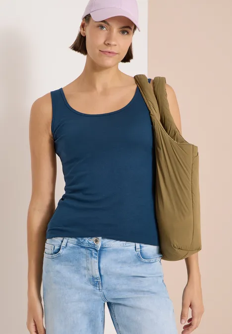 Femme en débardeur bleu et jean clair, sac olive à l'épaule, devant un fond neutre.