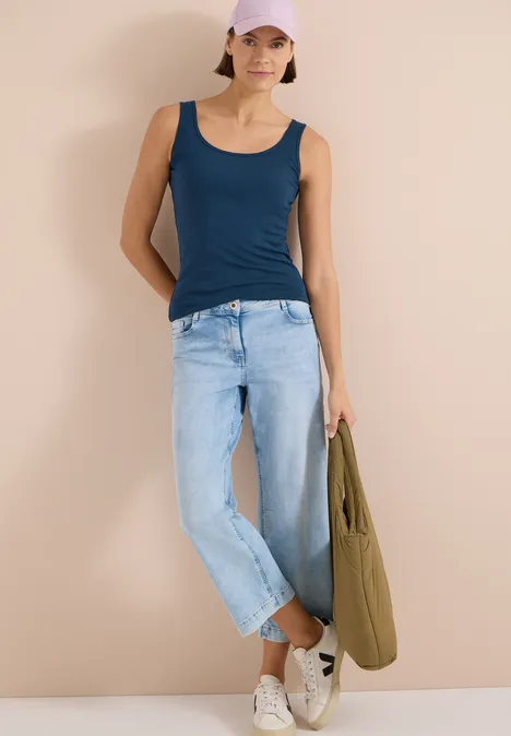 Une femme en débardeur bleu et jeans clair s'appuie sur un mur et tient un sac vert.