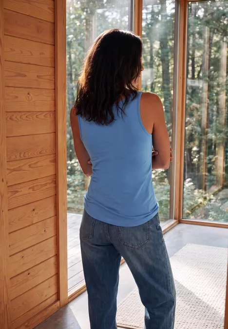 Basic Top aus reiner Baumwolle north blue Frau in blauem Top und Jeans steht in Holzhütte und blickt durch große Fenster in den Wald.