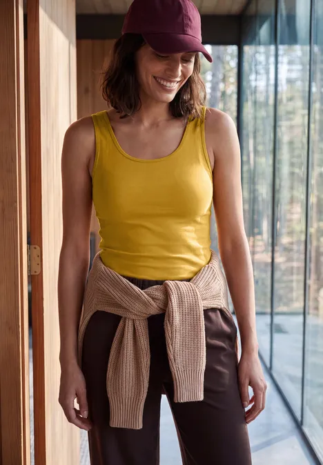 Vrouw met geel topje, bruine broek en pet lacht in modern huis, trui om haar middel gestrikt.