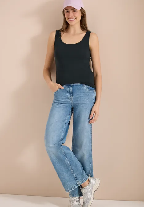 Frau trägt schwarzes Top, weite blaue Jeans und weiße Sneaker vor neutralem Hintergrund.