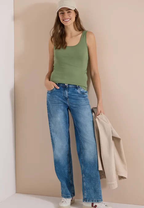 Vrouw in groen topje en blauwe jeans leunt tegen een beige muur, houdt een beige jas vast.