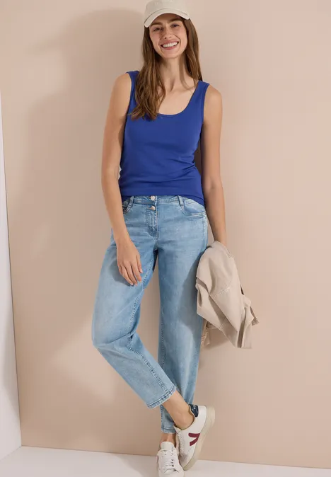 Vrouw leunt tegen lichte muur, draagt blauw topje, spijkerbroek, witte sneakers en houdt beige jas vast.