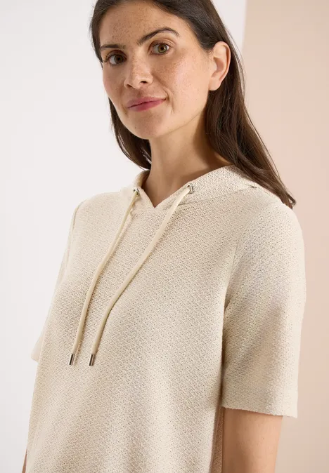 Vrouw met crèmegekleurd, kortmouwig gebreid hoodie met trekkoord.