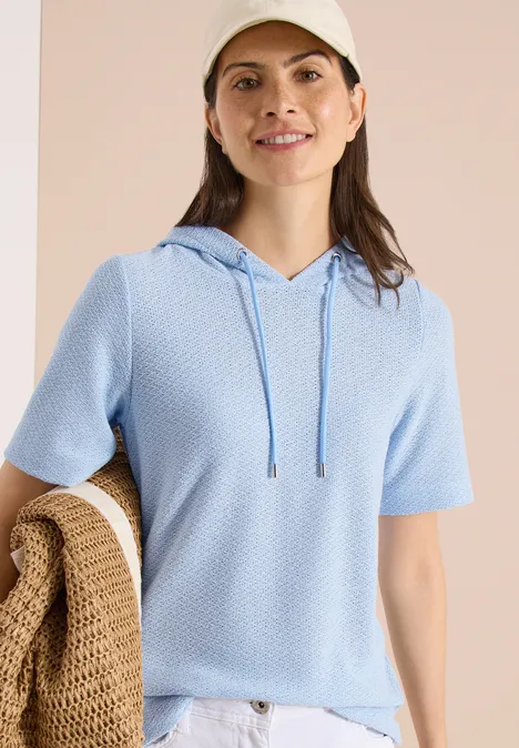 Vrouw draagt lichtblauw kort mouw capuchonshirt van fijngebreide stof, houdt een beige gehaakte tas vast.