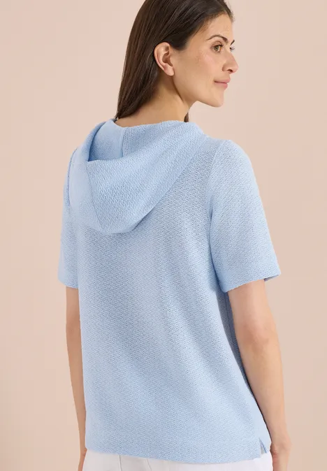 Achteraanzicht van een vrouw in lichtblauwe gebreide korte mouw hoodie voor een beige achtergrond.