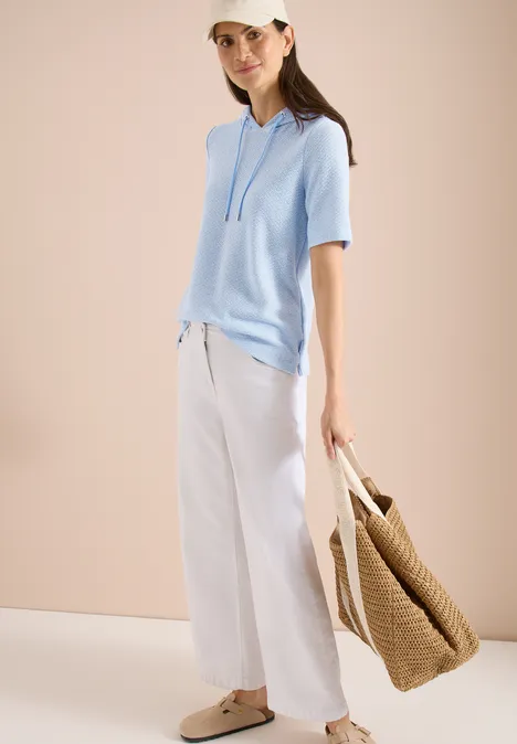 Persoon in lichtblauwe korte hoodie, witte wijde broek, strooien tas en slippers voor een neutrale achtergrond.