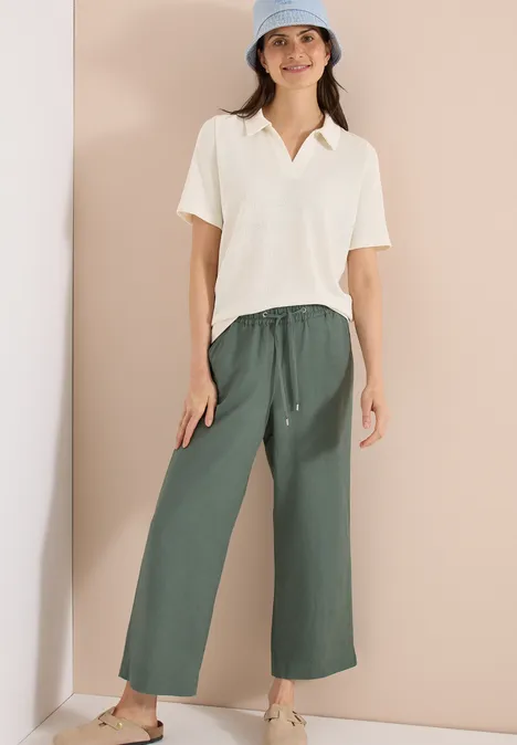 Vrouw in crème poloshirt en wijde groene stoffen broek voor effen achtergrond