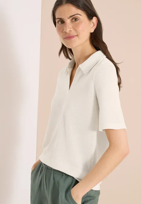 Vrouw in een witte polo met korte mouwen en een groene broek, zijwaarts staand met handen in de zakken.