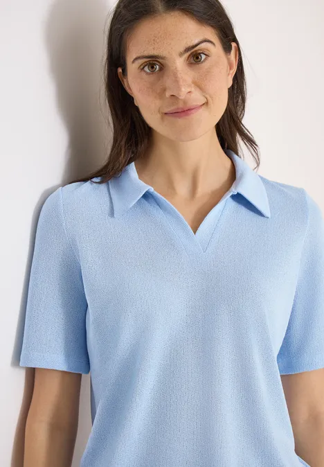 Vrouw in lichtblauw polohemd met korte mouwen voor een effen, lichte achtergrond.