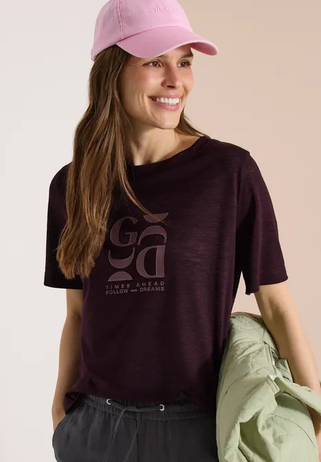 Eine Frau mit rosa Cap trägt lila T-Shirt und hält eine helle Jacke in der Hand.