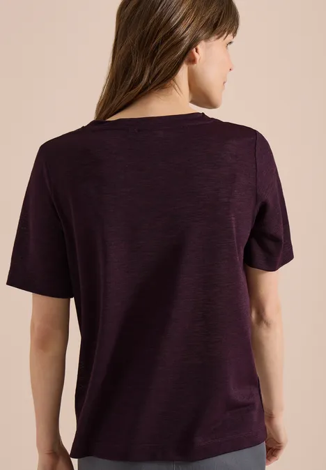 Frau von hinten in locker sitzender, dunkelvioletter Kurzarmshirt vor hellbeigem Hintergrund.