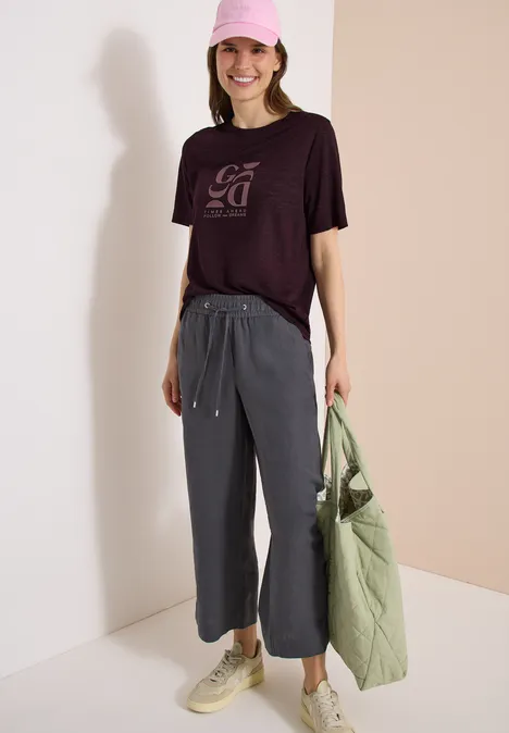 Frau in schwarzem T-Shirt und grauer Culottehose, helle Sneaker und grüne Stepp-Tasche, vor hellem Hintergrund.