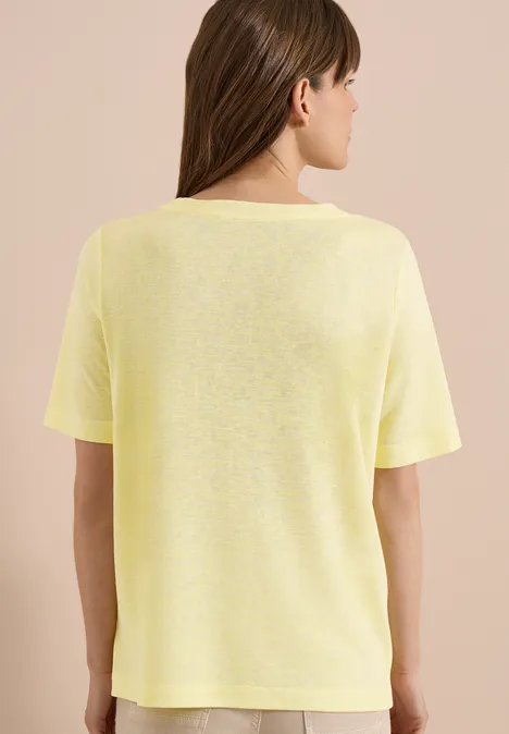 Frau trägt schlichtes gelbes T-Shirt, steht mit dem Rücken zur Kamera vor beigem Hintergrund.