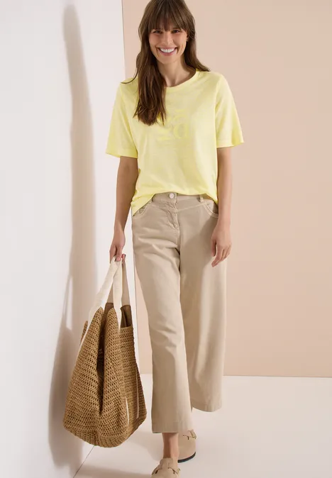 Frau in gelbem T-Shirt, beiger Hose mit großer Korbtasche vor neutralem Hintergrund.