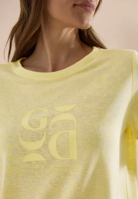 Frau trägt hellgelbes T-Shirt mit rundem Kragen und dezentem, spiegelverkehrtem Logo auf der Brust.