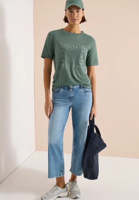 Persoon in groen T-shirt, blauwe jeans en sneakers, draagt een tas.