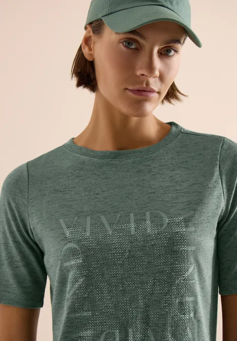 Persoon draagt groen T-shirt en pet voor lichte achtergrond, gezicht onscherp gemaakt.