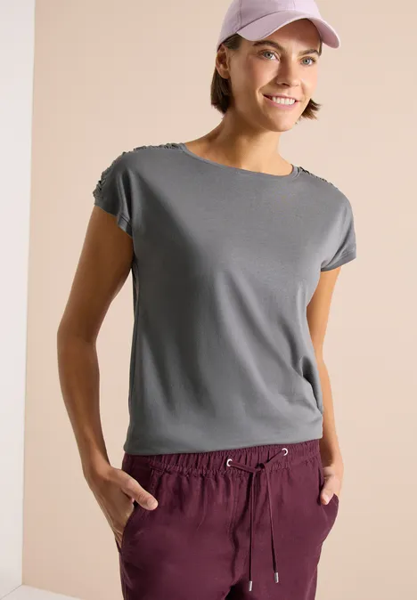 Frau trägt graues T-Shirt, weinrote Jogginghose und rosa Basecap vor neutralem Hintergrund.