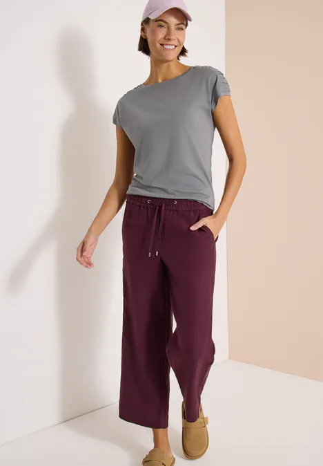 Person steht in einem zweifarbigen Eck, trägt graues T-Shirt, bordeauxrote Hose und Hausschuhe.