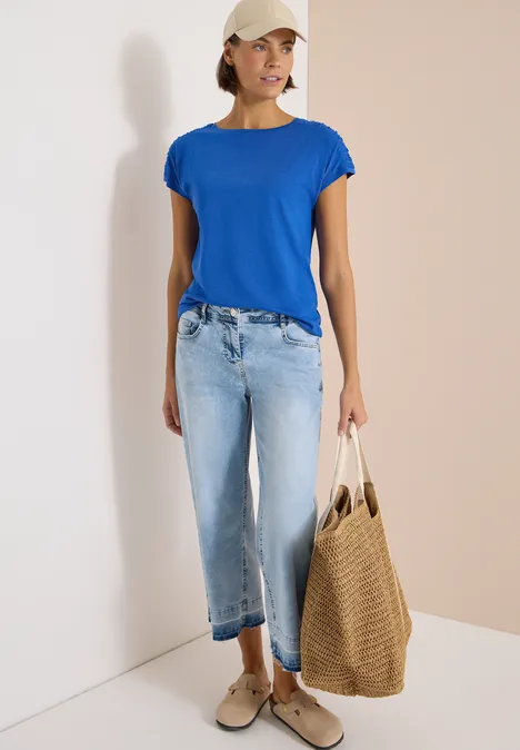 Kurzarm Shirt mit Boatneck und Raffung surf blue Frau steht in blauem T-Shirt, hellen Jeans, Sneakern und trägt eine große geflochtene Tasche.