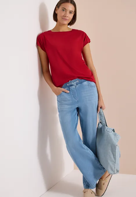 Kurzarm Shirt mit Boatneck und Raffung tamarillo red Frau lehnt an weißer Wand, trägt rotes Shirt, weite hellblaue Jeans und hellblaue Tasche.