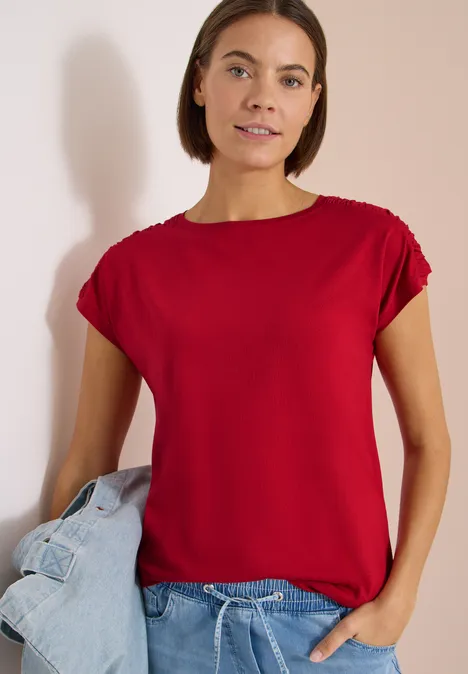 Kurzarm Shirt mit Boatneck und Raffung tamarillo red Eine Frau in rotem T-Shirt hält eine blaue Jeansjacke vor hellem Hintergrund.