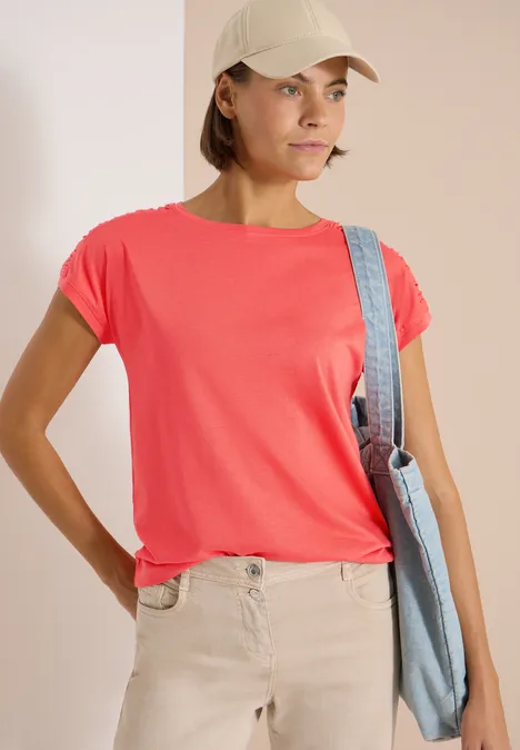 Kurzarm Shirt mit Boatneck und Raffung shiny coral Frau in rotem T-Shirt, beiger Hose und Cap, trägt eine Umhängetasche.