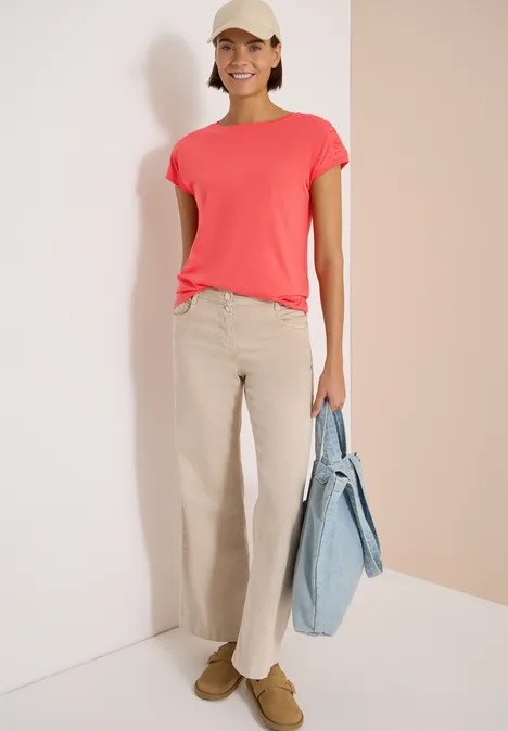 Kurzarm Shirt mit Boatneck und Raffung shiny coral Person trägt korallenrotes T-Shirt, beige Hose und hellblaue Tasche vor neutralem Hintergrund.