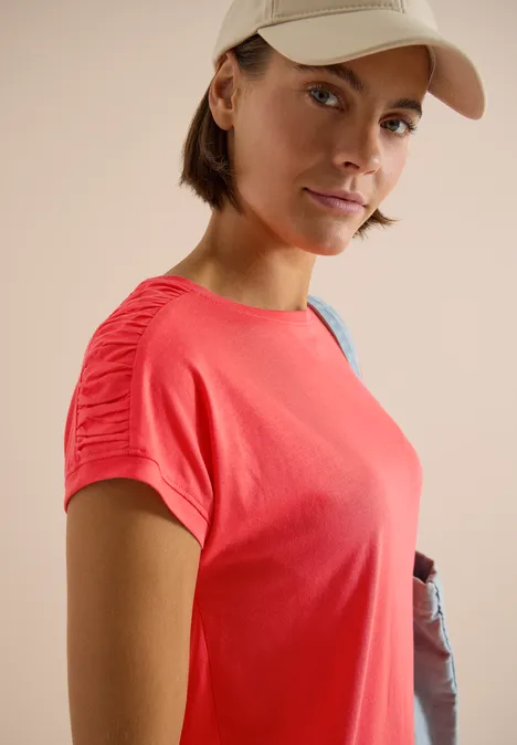 Kurzarm Shirt mit Boatneck und Raffung shiny coral Person in rotem T-Shirt und Baseballcap mit Umhängetasche vor neutralem Hintergrund.