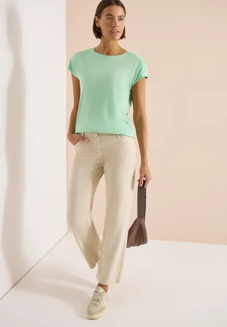 Eine Frau trägt ein mintgrünes T-Shirt, beigefarbene weite Hosen und eine Tasche, stehend in hellem Raum.