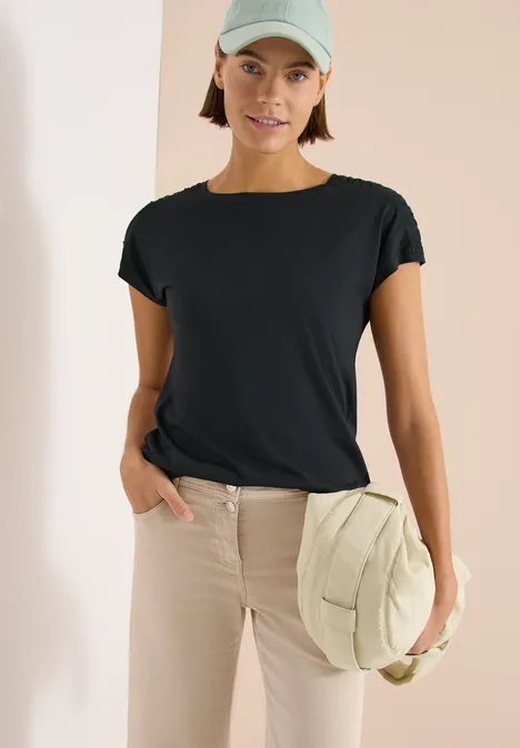 Persoon draagt een zwart T-shirt en een beige broek, houdt een ronde beige tas vast.