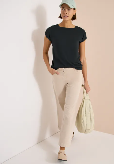 Vrouw leunt tegen lichte muur, draagt zwart shirt, wijde beige jeans, lichte schoenen en houdt een gewatteerde tas vast.