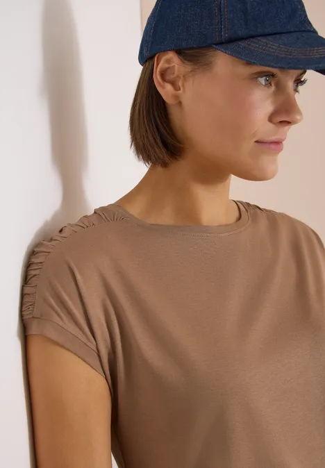 Kurzarm Shirt mit Boatneck und Raffung creamy mocha Eine Person trägt ein braunes T-Shirt mit gerafften Ärmeln und eine dunkelblaue Baseballkappe.