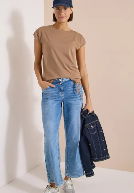 Kurzarm Shirt mit Boatneck und Raffung creamy mocha Eine Frau trägt ein beiges T-Shirt, blaue Jeans und hält eine Jeansjacke in der Hand.