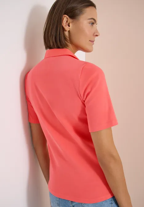 Rückenansicht einer Person in korallenpinken Poloshirt mit kurzen Ärmeln, die entspannt an einer Wand lehnt.
