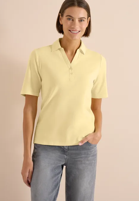 Basic Poloshirt mit Kurzarm buttermilk yellow Frau in gelbem Poloshirt und Jeans steht vor hellem Hintergrund, eine Hand in der Hosentasche.