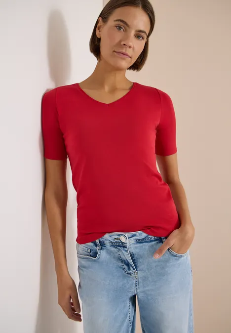 Eine Frau in rotem T-Shirt und blauen Jeans steht entspannt mit einer Hand in der Tasche vor hellem Hintergrund.