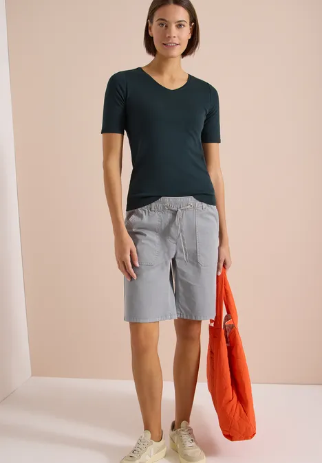 Eine Person trägt ein dunkelgrünes T-Shirt, graue Bermudashorts und hält eine orangefarbene Tasche.