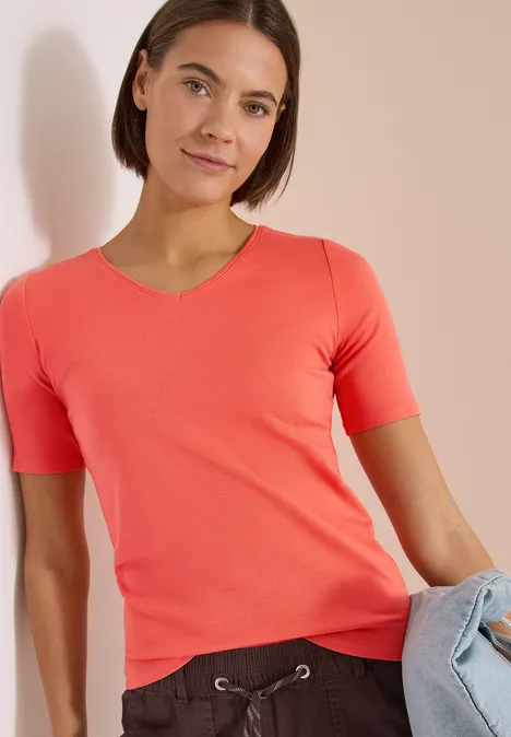 Model in korallenfarbenem T-Shirt hält eine hellblaue Jeans vor neutralem Hintergrund.