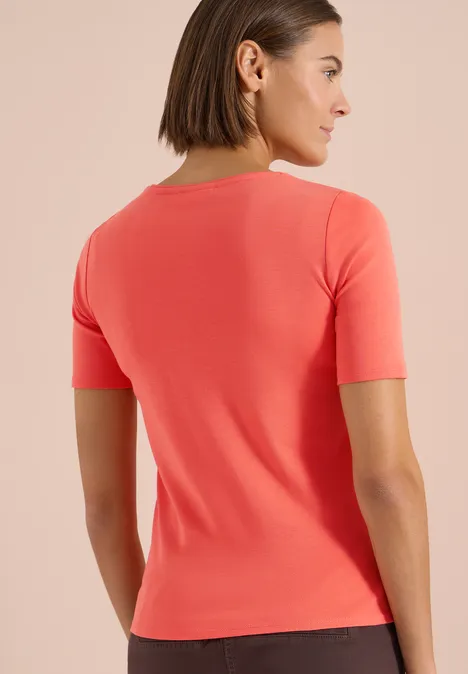 Rückenansicht einer Frau in enganliegendem, korallrotem Kurzarmshirt vor neutralem Hintergrund.
