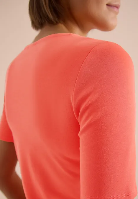 Rückenansicht einer Person in korallenfarbenem T-Shirt mit kurzen Ärmeln vor hellem Hintergrund.