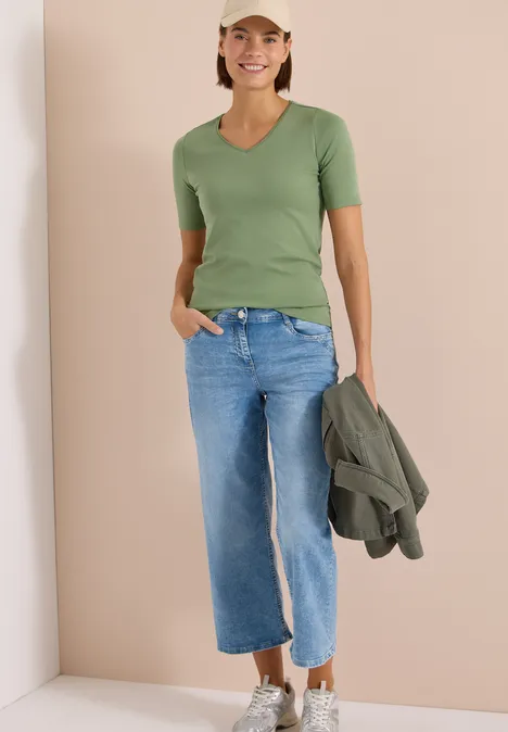 Basic T-Shirt aus Baumwolle deep matcha green Person vor beigem Hintergrund trägt grünes T-Shirt, weite blaue Jeans und hält eine grüne Jacke.