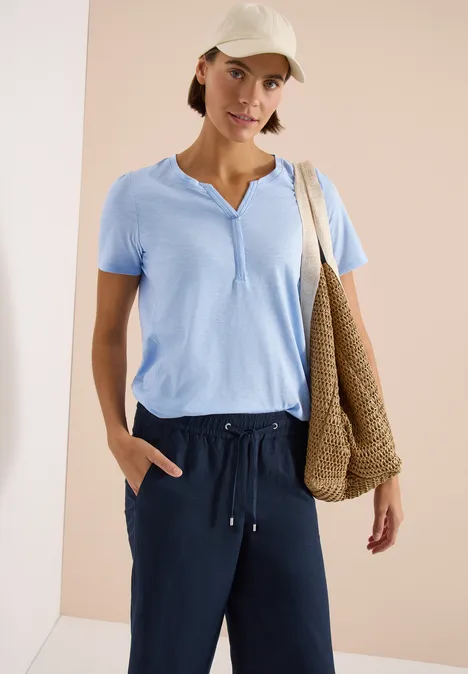 Frau trägt hellblaues Kurzarmshirt, dunkelblaue Hose, Cap und geflochtene Strohtasche vor neutralem Hintergrund.