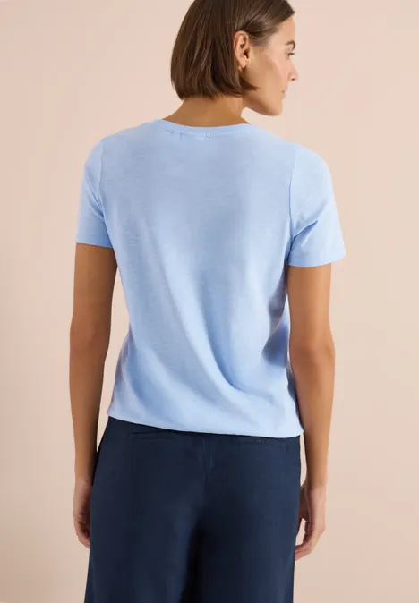 Rückansicht einer Person in hellblauem T-Shirt und dunkelblauer Hose vor beigem Hintergrund.